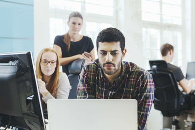 Businesswomen looking at male colleague using laptop at desk in creative office