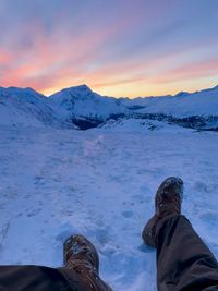 Low section of man relaxing on snowcapped mountains against sky during sunset