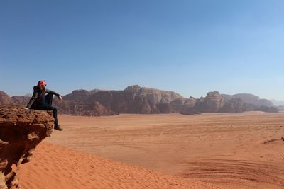 Side view of man sitting on cliff by arid landscape against clear sky