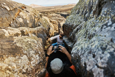 Anonymous traveler shooting friend via smartphone while standing in narrow crack between rough rocks in countryside of fuerteventura, spain