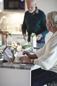 Senior woman using laptop while male partner standing by kitchen island