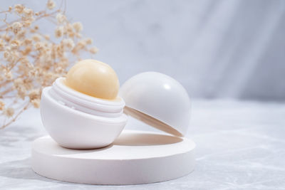 Round light colored lip balm in white plastic case on podium with flowers, mockup design, label