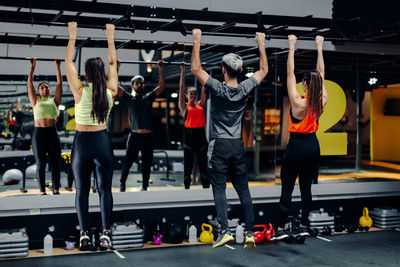 Full body back view of athletic man and women in sportswear hanging on sports bar while doing abs exercise together in front of mirror in contemporary gym