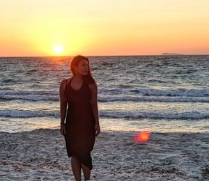 Woman looking away while standing at beach against sky during sunset