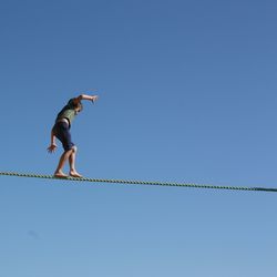 Low angle view of boy walking on tightrope against clear blue sky