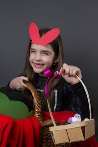 Portrait of smiling girl with costume rabbit ears sitting in basket against gray background