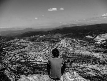 Rear view of man sitting on mountain looking at landscape against sky