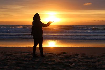 Silhouette woman standing on shore at beach against orange sky during sunset