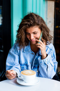 Charming woman with beautiful smile talking by mobile phone during rest, drinking cappuccino