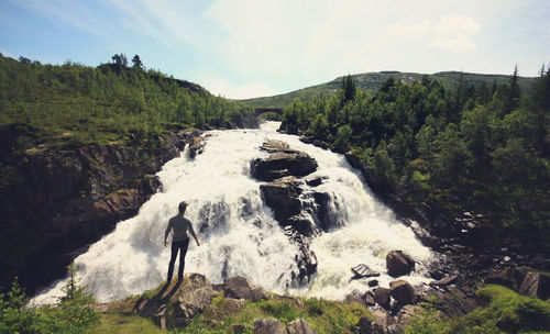 Full length rear view of man standing by waterfall against sky