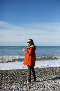 Young woman in cold weather on the seashore uses a mobile phone