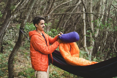 A man packs a sleeping bag and hammock at campsite in the forest