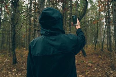 Rear view of person wearing hood photographing trees in forest during autumn