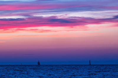 Dramatic purple and pink sky at dusk over ocean with sailing vessel silhouetted in background 