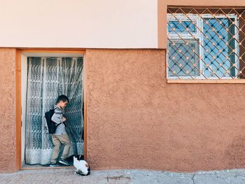 FULL LENGTH OF YOUNG MAN SITTING AGAINST WINDOW ON BUILDING