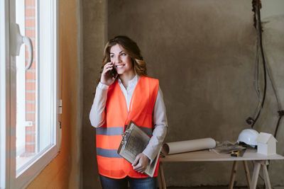 Professional architect woman in construction site talking on mobile phone holding blueprints