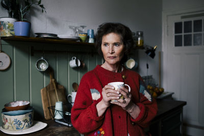 Thoughtful lonely retired woman holding mug while standing in kitchen at home