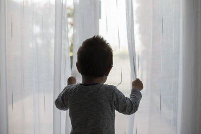 Rear view of boy standing by curtain at home