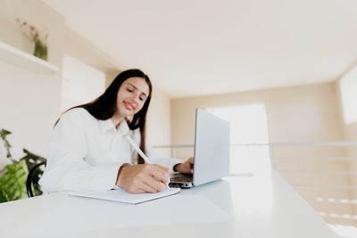 Caucasian young business woman sitting at desk, reading from computer, taking notes at notebook
