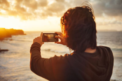 Rear view of man photographing sea during sunset