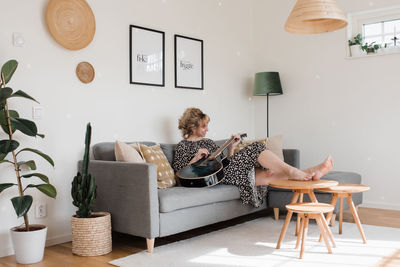 Woman sat on the couch in a dress playing the guitar at home relaxing