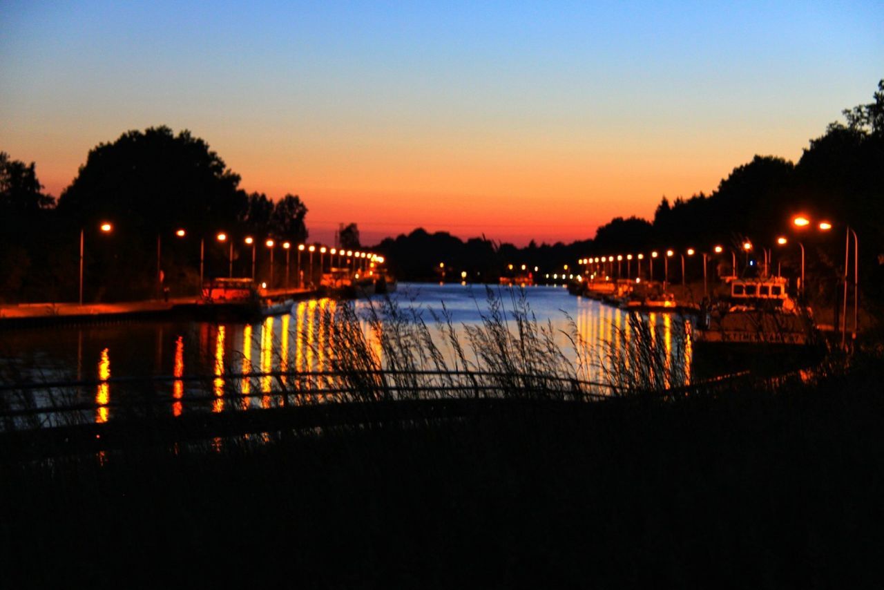 illuminated, water, sunset, reflection, silhouette, night, clear sky, copy space, tranquil scene, dusk, tranquility, river, lake, scenics, orange color, sky, nature, street light, waterfront, beauty in nature