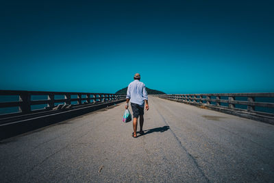 Rear view of man walking on bridge against clear blue sky