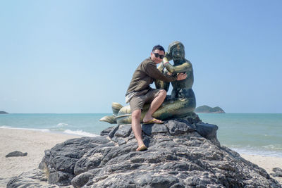 Portrait of woman sitting with mermaid statue at beach against blue sky