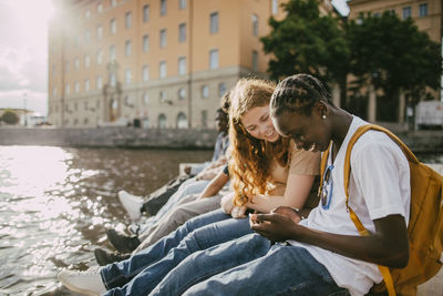 Teenage boy using smart phone while sitting with female friend near lake at sunny day