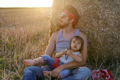 Father and son in t-shirts sitting next to a haystack on a sloping field during sunset