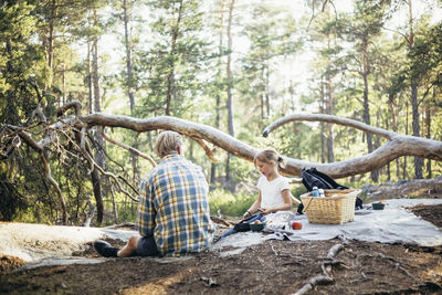 Daughter sitting with father on picnic blanket and using phone in forest