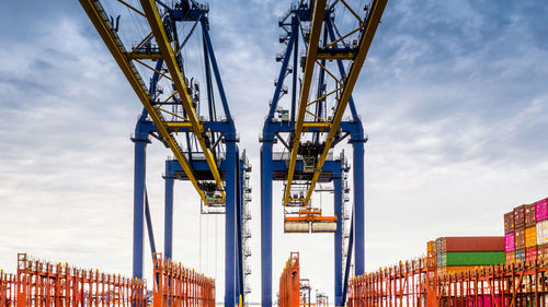 Low angle view of cranes and cargo containers against cloudy sky