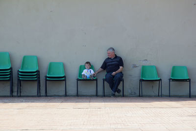 Senior man sitting with boy on chairs against wall