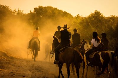 People riding horses on field against sky during sunset