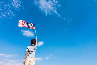 Rear view of boy holding malaysian flag while standing against blue sky
