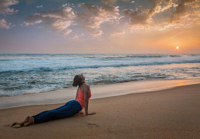 Woman sitting on shore at beach against sky during sunset
