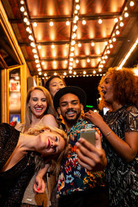 Young content diverse partners making faces while taking self portrait on cellphone under shiny lights during night celebration