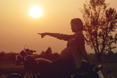 Pregnant woman pointing while sitting on motorcycle against sky during sunset