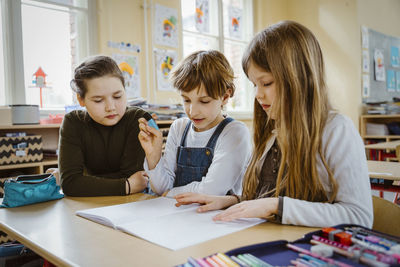 Boy and girls sharing book while studying together at desk in classroom