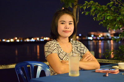 Portrait of young woman with arms crossed sitting against sea at outdoor cafe during night