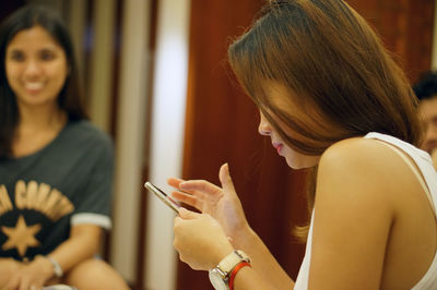 Side view of young woman using phone with friend in background