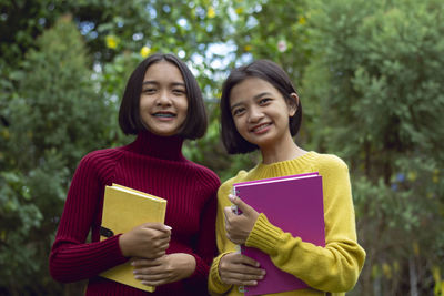 Portrait of smiling teenage girl with friend holding books against trees