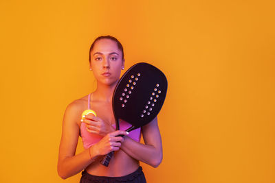 Portrait of woman holding racket and tennis ball against orange background