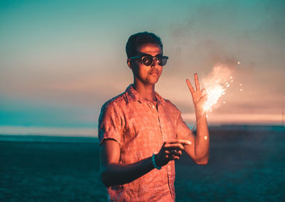 Young man wearing sunglasses while holding sparkler against sky during sunset
