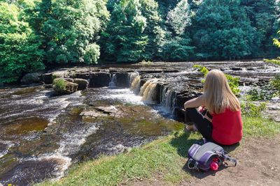 Rear view of woman sitting on grass by stream against trees