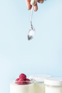 Cropped hand of woman holding spoon over raspberries and yogurt in drinking glass against blue background