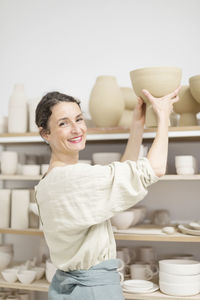 Portrait of smiling mid adult potter holding ceramics in workshop
