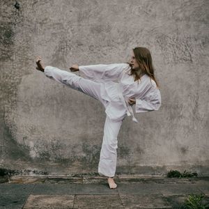 Full length of young woman wearing white uniform while practicing karate against wall