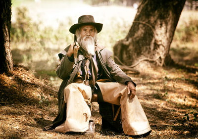 Man holding gun while sitting on grass by tree