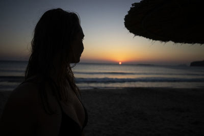 Rear view of woman standing at beach against sky during sunset